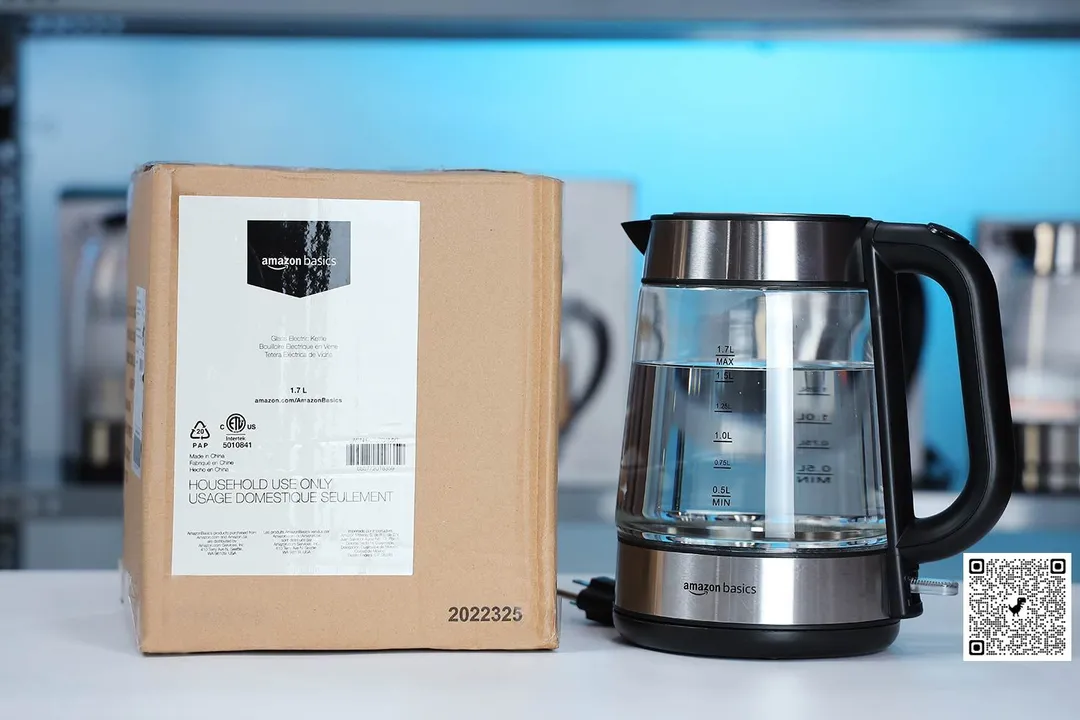 The Amazon Basics Electric Kettle (F-625C) on the right and its cardboard box on the left. In the background is a shelf with different electric kettles.