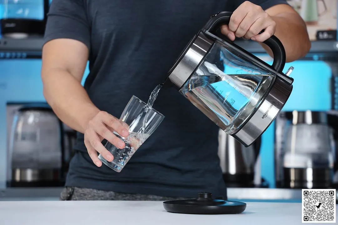 A person in a dark green shirt pouring water from the Amazon Basics Electric Glass and Steel Kettle (F-625C) into a glass.