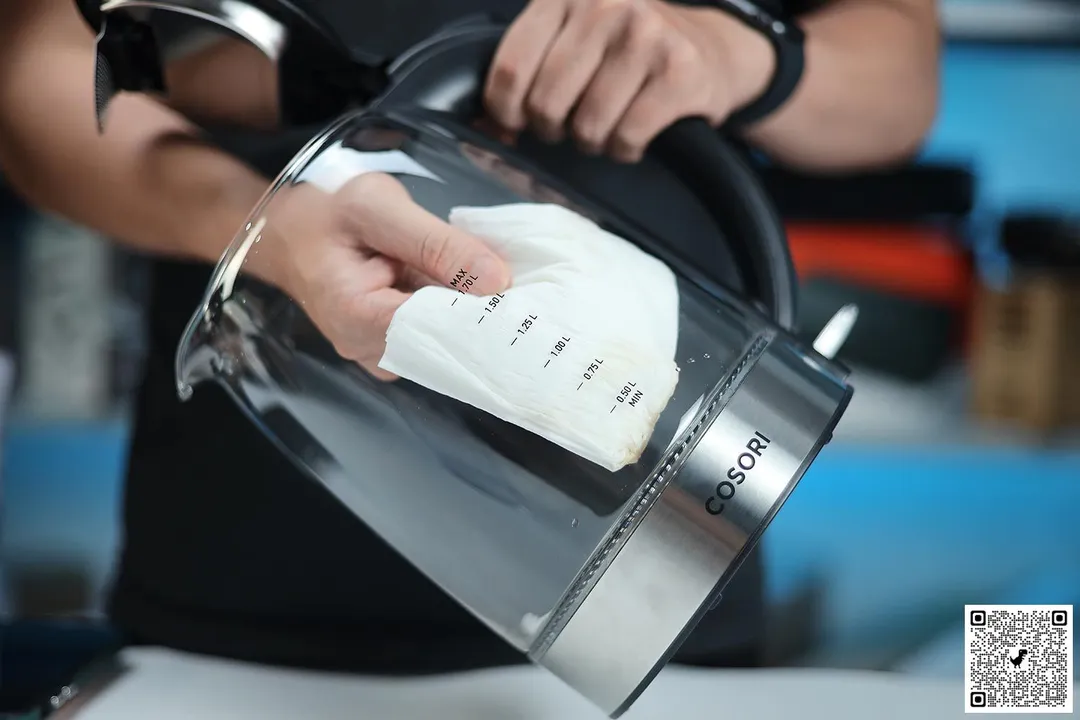 A person in a black shirt holding the Cosori Glass Electric Kettle (GK172-CO) by its handle on one hand and the other hand wiping the carafe interior with a piece of tissue.