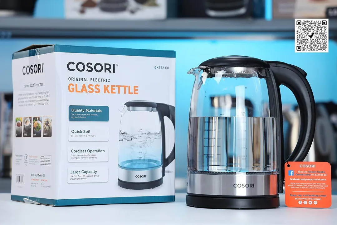 The Cosori Glass Electric Kettle (GK172-CO) on the right and its cardboard box on the left. In the background is a shelf with different electric kettles.