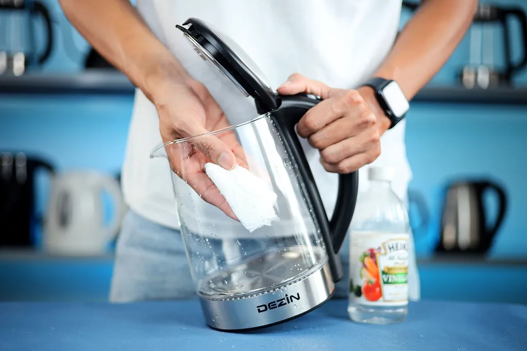 A person in a white shirt holding the Zeppoli Electric Kettle ZPL-KETTLE by its handle on one hand and the other hand wiping the carafe interior with a piece of tissue. On the right is a bottle of white vinegar.