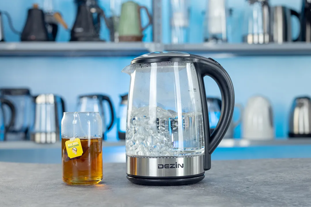The Dezin Electric Glass Kettle DZ380 on the right and a cup of black tea on the left. In the background is a shelf with different electric kettles.