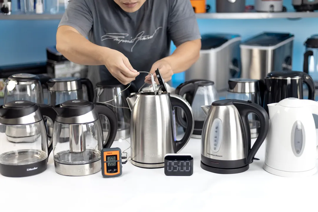 Man using a thermometer agaisnt an electric kettle on the table, surrounded by other electric kettles and a digital timer