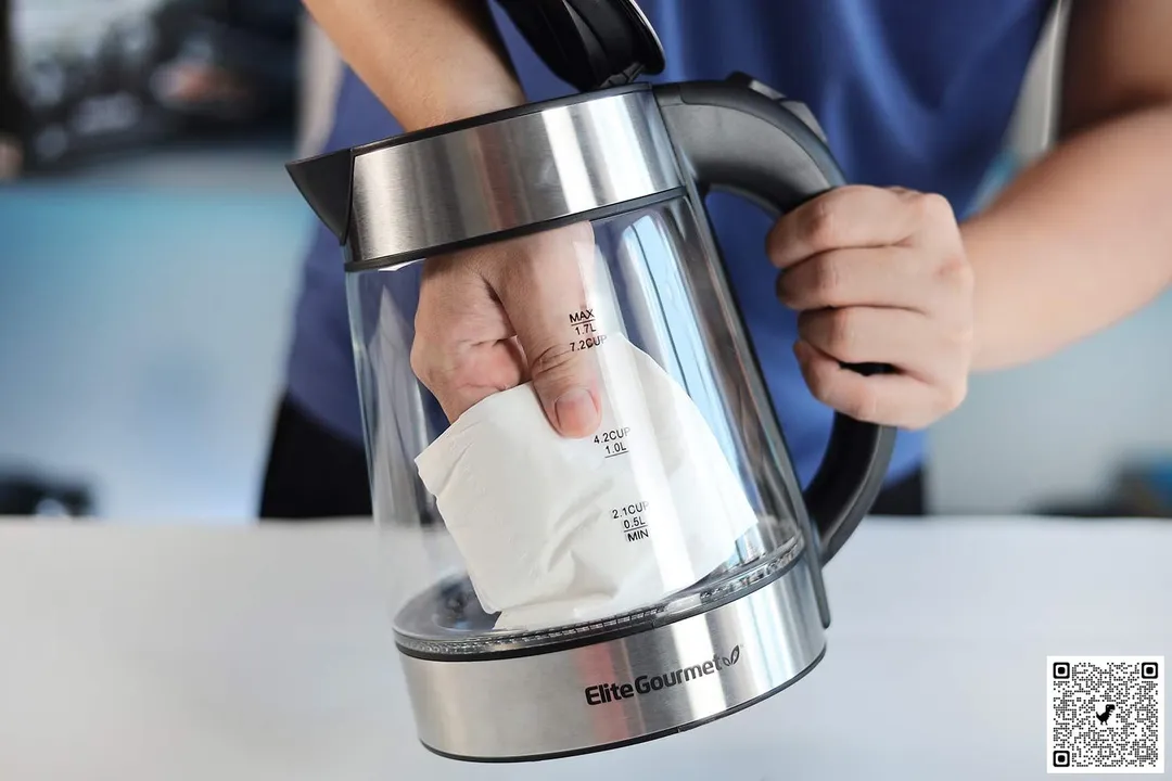 A person in a blue shirt holding the Elite Gourmet Electric Glass Kettle (EKT-602) by its handle on one hand and the other hand wiping the carafe interior with a piece of tissue.