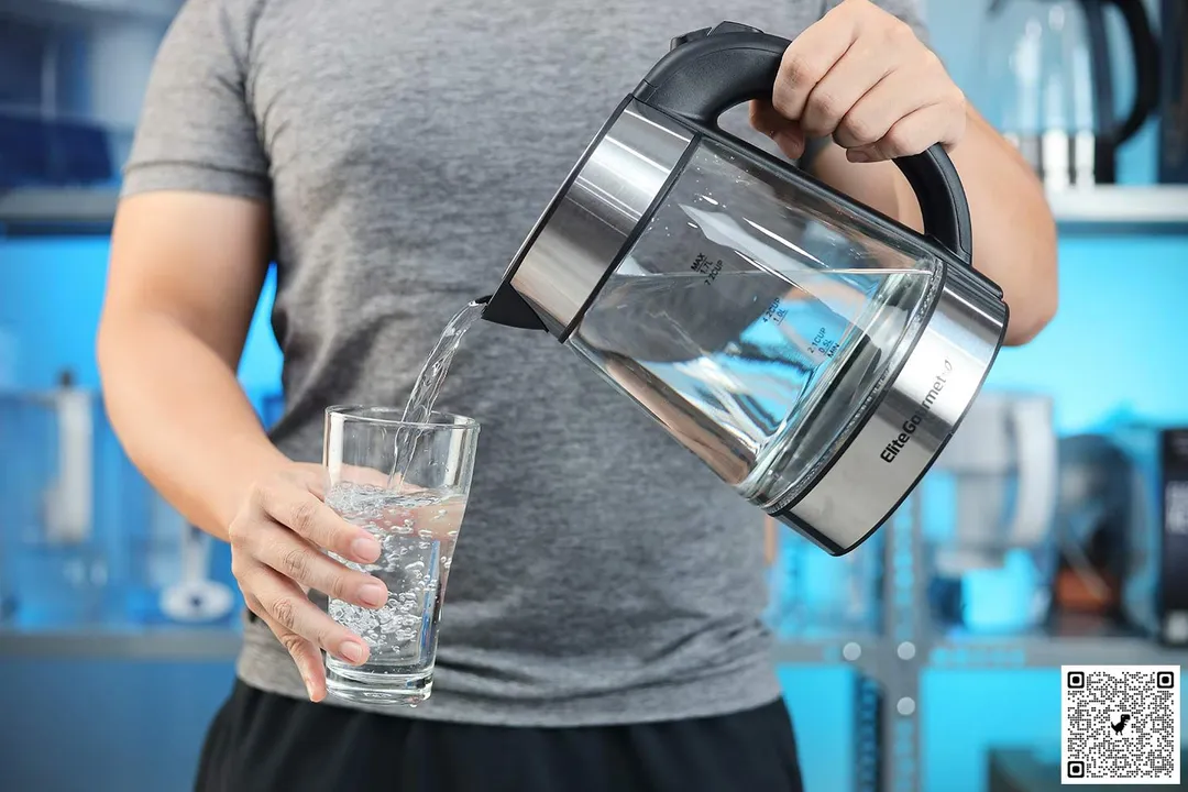 A person in a grey shirt pouring water from the Elite Gourmet Electric Glass Kettle (EKT-602) into a glass.