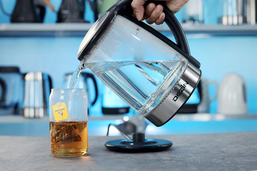A hand pouring water from the Dezin Electric Glass Kettle DZ380 into a glass of black tea