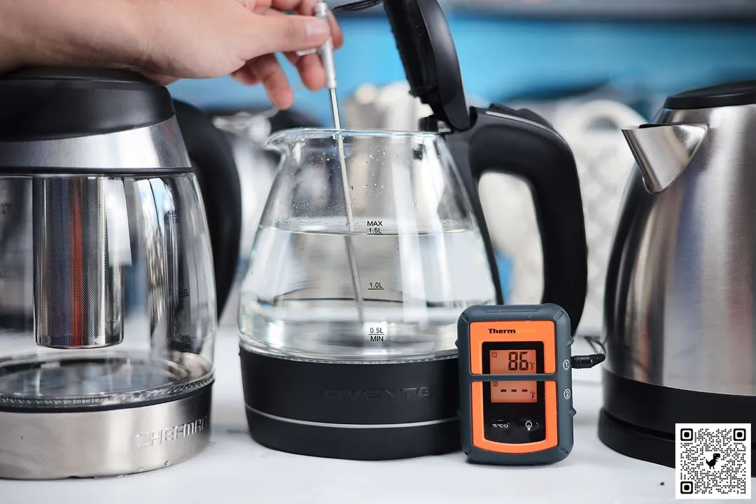 In the foreground, on a white table are 3 electric kettles. The kettle in the middle is filled with 1.5 liters of water, a hand is holding a probe inside it. The orange ThermoPro digital thermometer displays the water temperature to be 86°F.