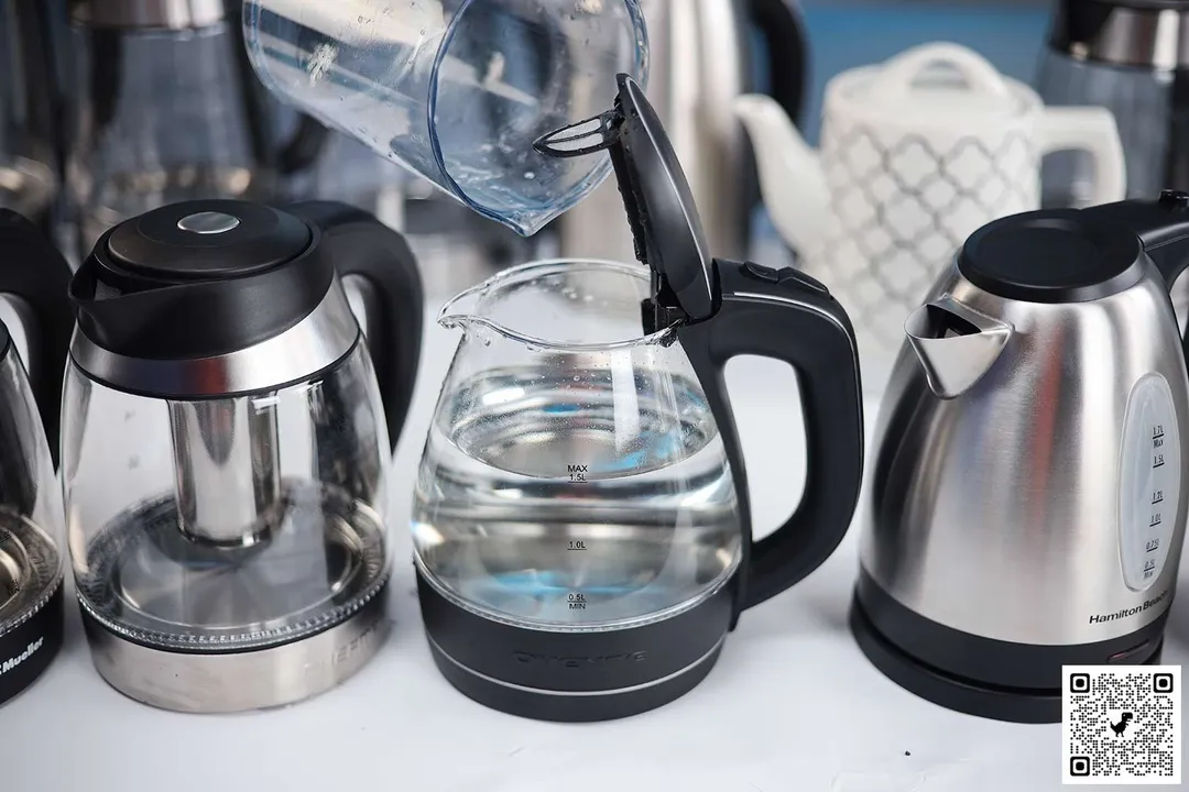 In the foreground, on a white table are 3 electric kettles. The kettle in the middle is filled with 1.5 liters of water from the measuring cup above. In the background are more electric kettles.