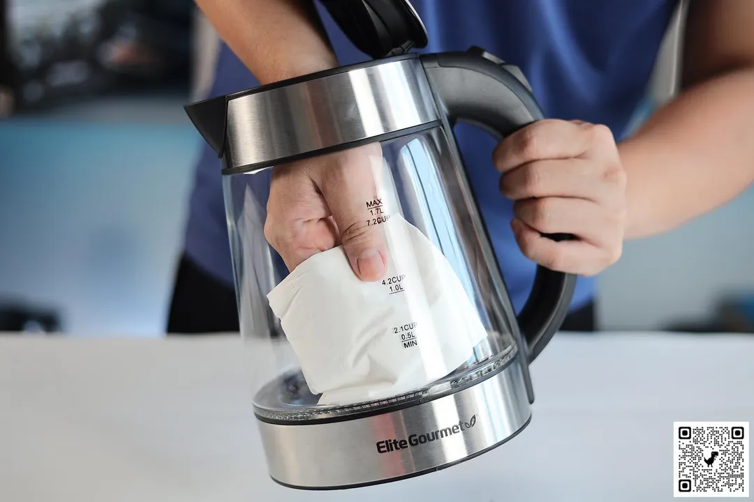 A person in a blue shirt holding the Elite Gourmet Electric Glass Kettle EKT-602 by its handle on one hand and the other hand wiping the carafe interior with a piece of tissue.