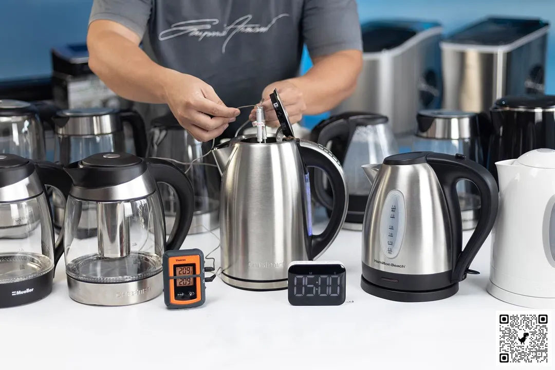 There are 9 electric kettles on a white table. A person in a grey shirt holding two probes inside the stainless steel electric kettle in the middle. On the left of the kettle is a Thermo Pro two-probe digital thermometer displaying 209°F for both probes. On the right of the kettle is a digital timer displaying 5 minutes on the countdown. In the background is a shelf of ice-makers.