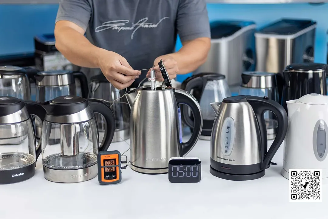 There are 9 electric kettles on a white table. A person in a grey shirt holding two probes inside the stainless steel electric kettle in the middle. On the left of the kettle is a Thermo Pro two-probe digital thermometer displaying 209°F for both probes. On the right of the kettle is a digital timer displaying 5 minutes on the countdown. In the background is a shelf of ice-makers.