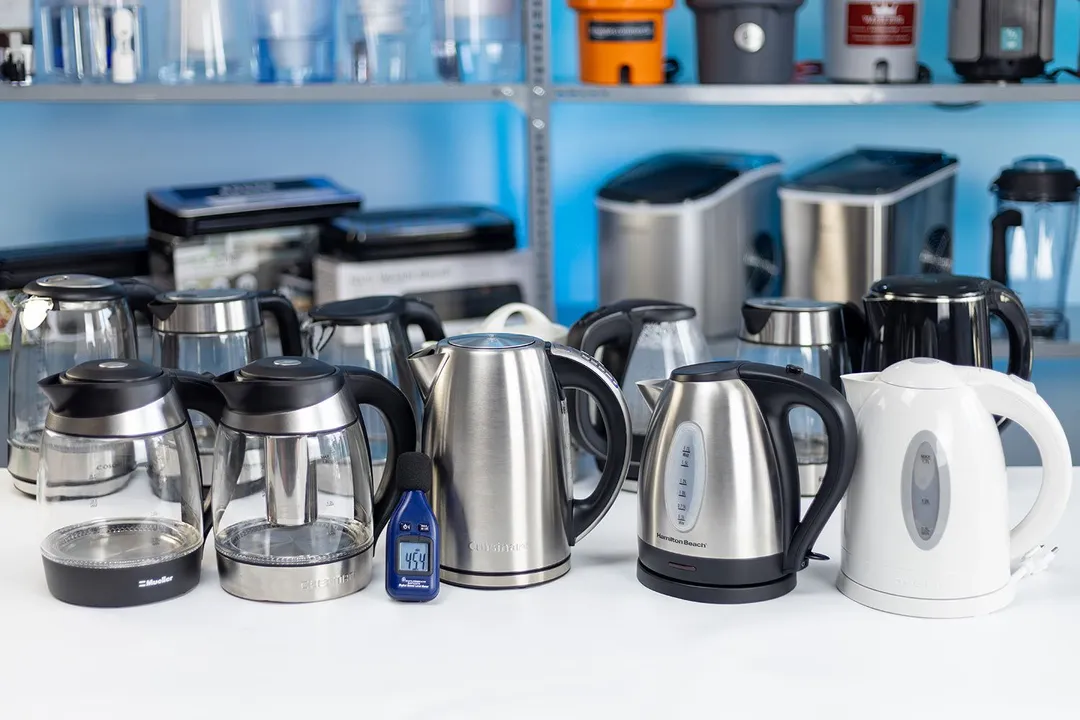 On a white table is 12 electric kettles. In front of them is a blue noise meter. In the background is a shelf of ice-makers.
