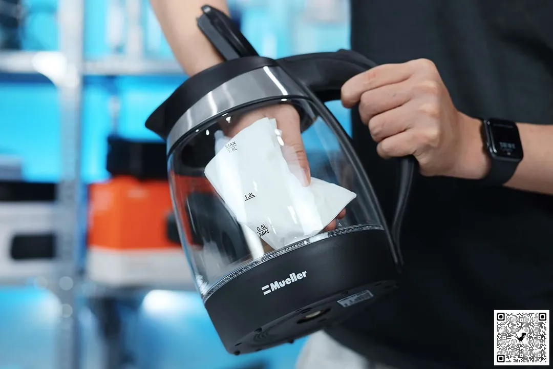 A person in a black shirt holding the Mueller Ultra Electric Kettle (M99S) by its handle on one hand and the other hand wiping the carafe interior with a piece of tissue.