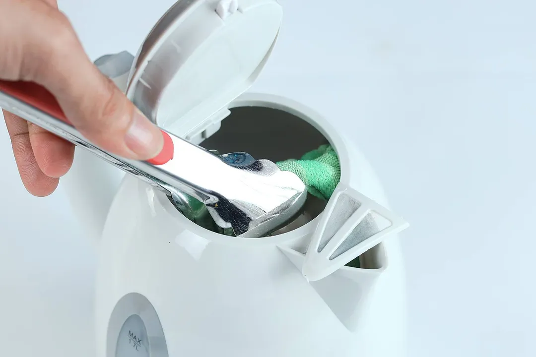 A hand using a kitchen tong to wipe the interior of the Ovente Electric Kettle KP72W with a soft green cloth.