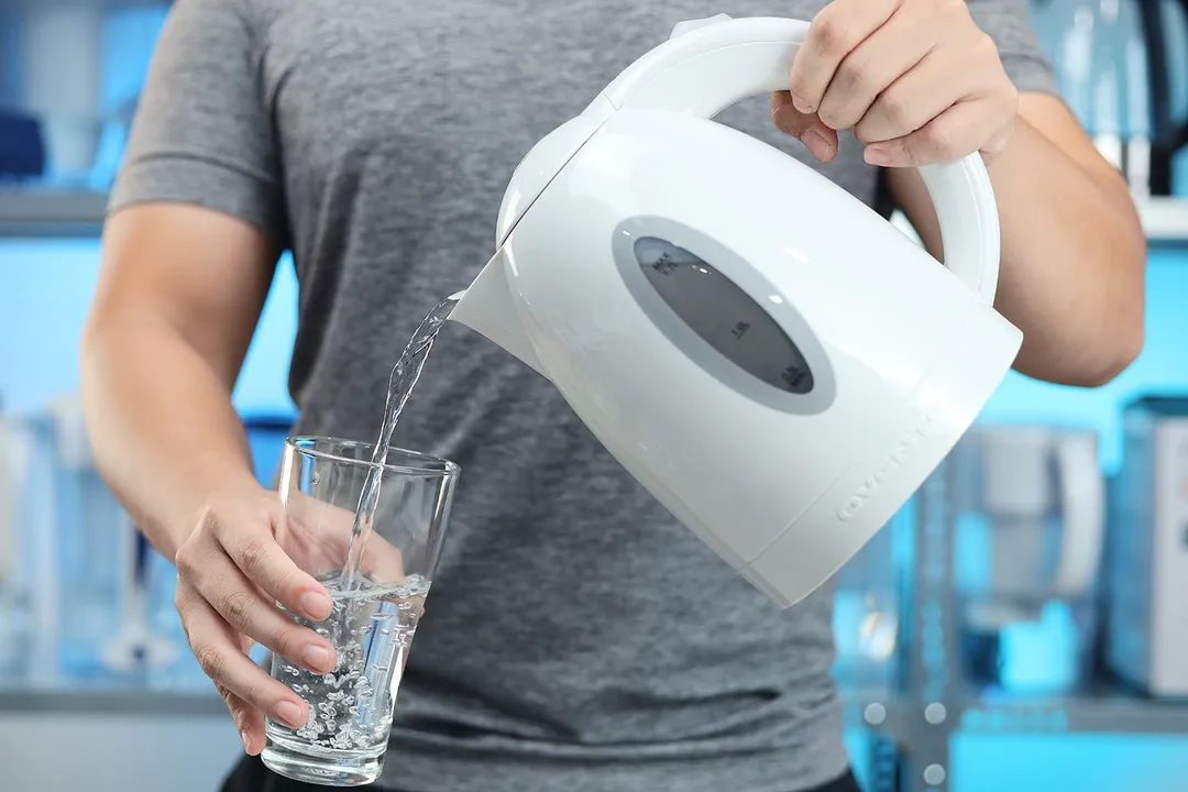 A person in a grey shirt pouring water from the Ovente Electric Kettle KP72W  into a glass.