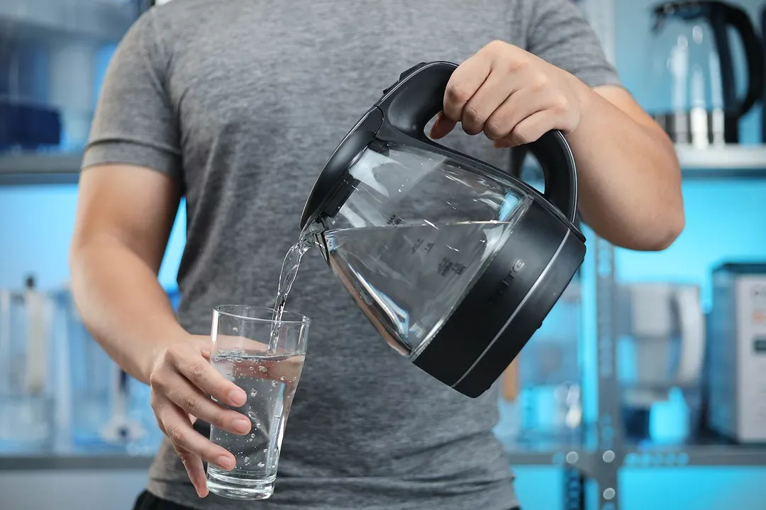 A person in a grey shirt pouring water from the Ovente Electric Glass Kettle (KG83B) into a glass.