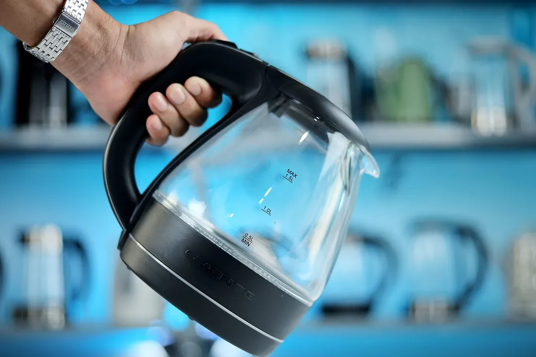A hand holding the Ovente Electric Glass Kettle (KG83B) by its handle.
