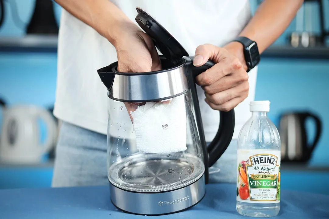  A person in a white shirt holding the Zeppoli Electric Kettle ZPL-KETTLE by its handle on one hand and the other hand wiping the carafe interior with a piece of tissue. On the right is a bottle of white vinegar.