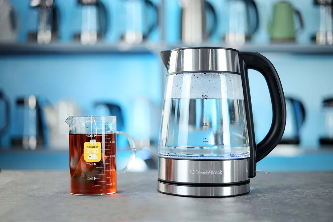 The Peach Street Electric Kettle PE-1300 on the right and a cup of black tea on the left. In the background is a shelf with different electric kettles.