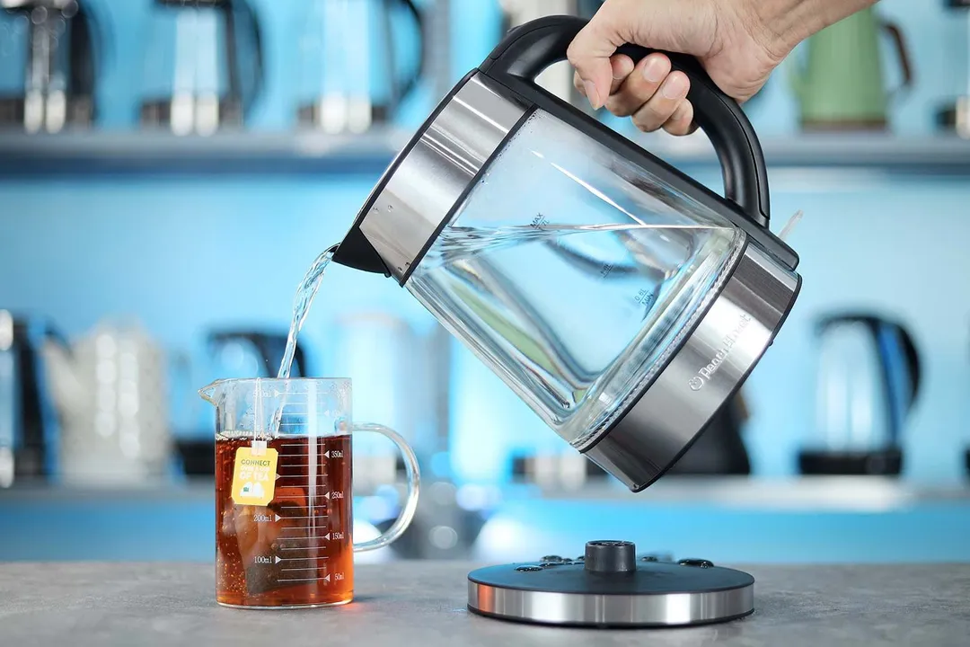A hand pouring water from the Peach Street Electric Kettle PE-1300 into a glass of black tea.