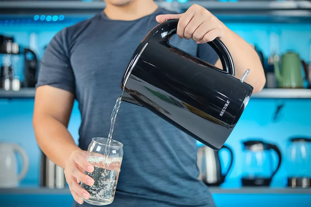 A person in a grey shirt pouring water from the Secura Electric Stainless Steel Double-Wall Kettle (SWK-1701DA) into a glass.