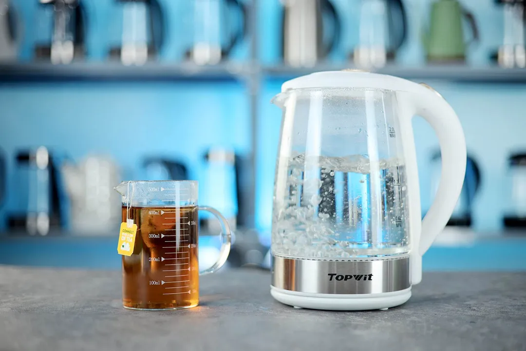 The Topwit Glass Electric Tea Kettle T630 on the right and a cup of black tea on the left. In the background is a shelf with different electric kettles.