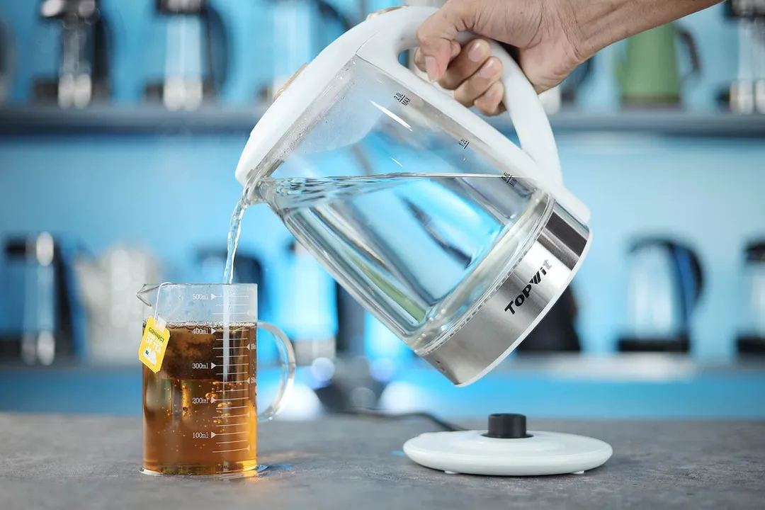 A hand pouring water from the Topwit Glass Electric Tea Kettle T630 into a glass of black tea.