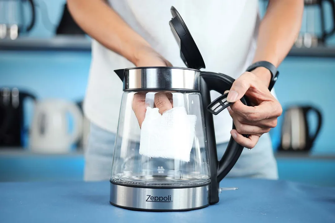 A person in a white shirt holding the Zeppoli Electric Kettle ZPL-KETTLE by its handle on one hand and the other hand wiping the carafe interior with a piece of tissue.