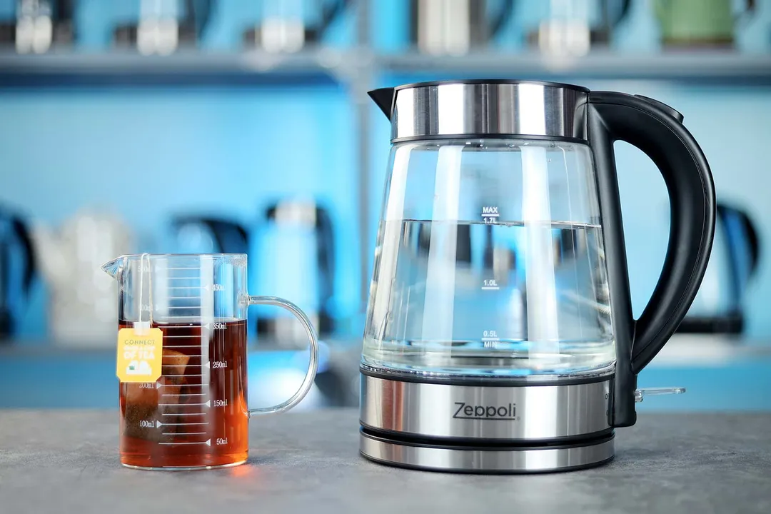 The Zeppoli Electric Kettle ZPL-KETTLE on the right and a cup of black tea on the left. In the background is a shelf with different electric kettles.