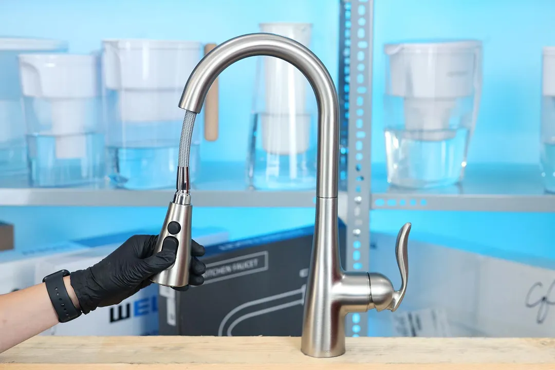 A hand pulls on the sprayer of a Moen Arbor 7594SRS kitchen faucet installed on a wood platform.