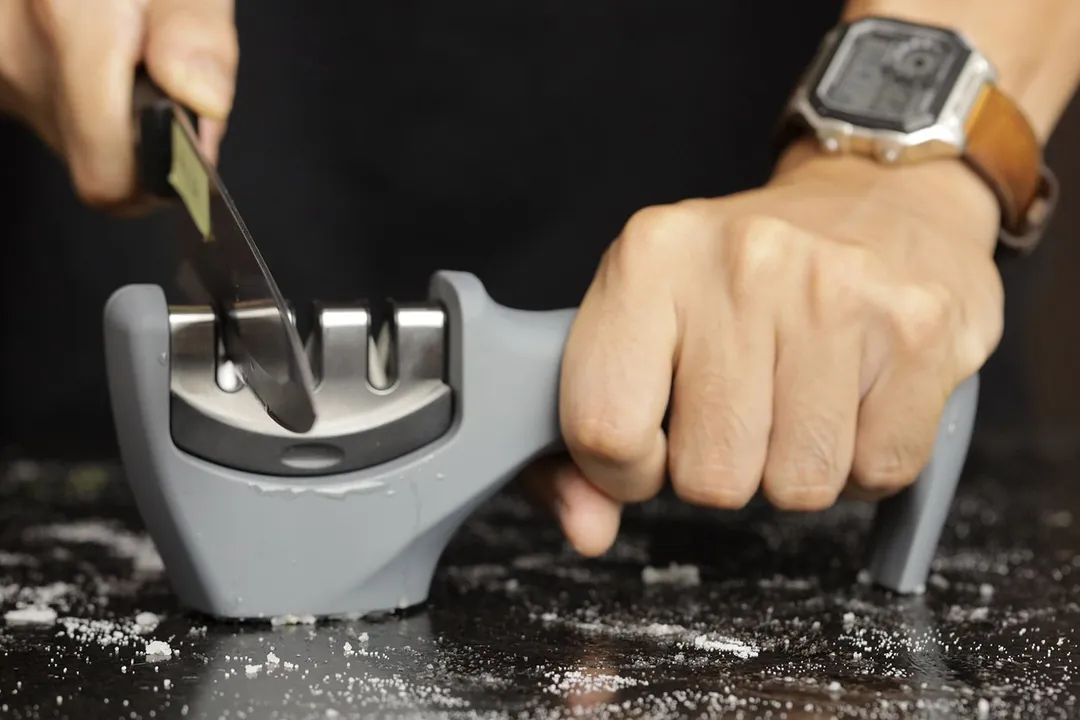 Two hands holding and sharpening a kitchen knife with the Amesser 3-slot knife sharpener on a salt-sprinkled countertop