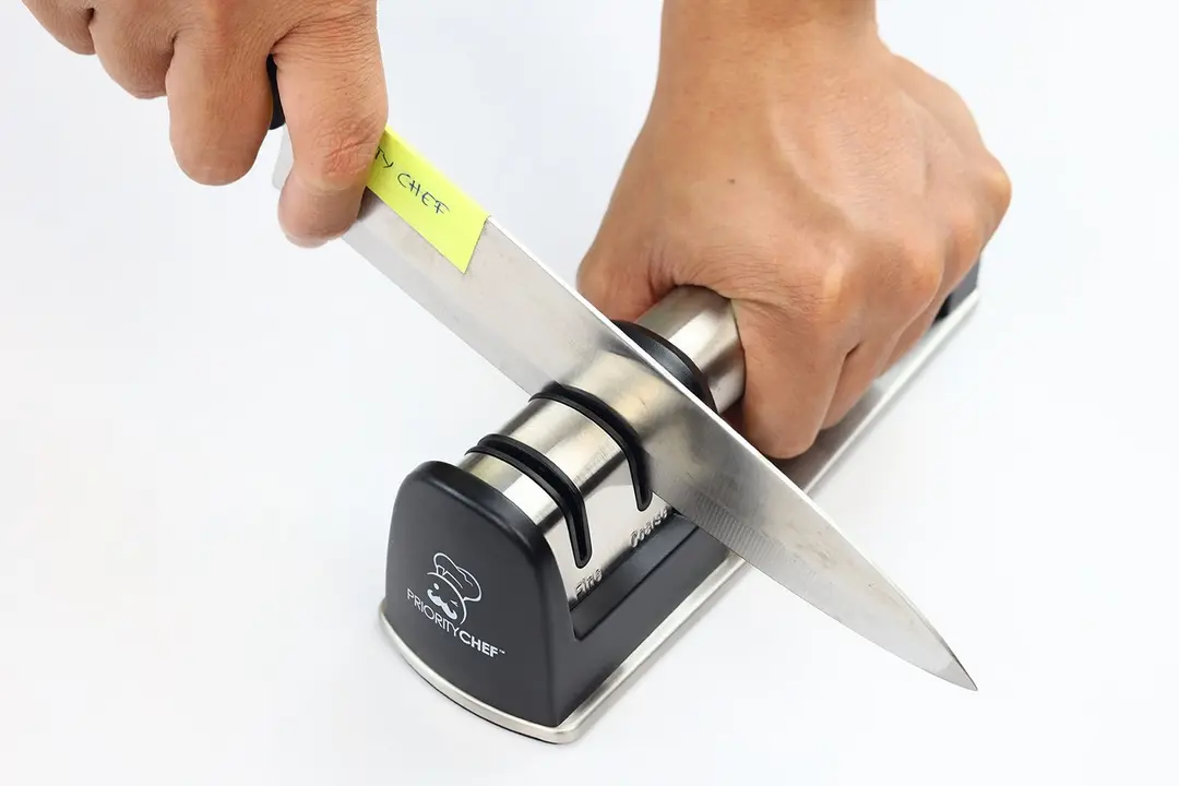 A kitchen knife being sharpened with the Priority Chef, and the two hands that hold the knife and the sharpener