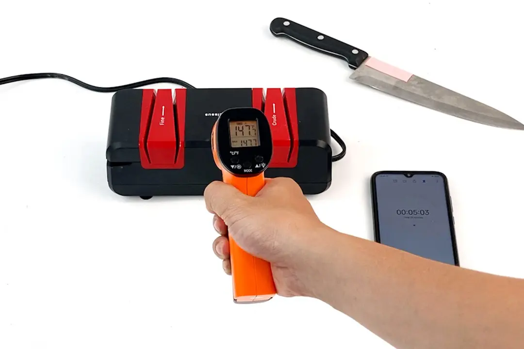 A hand holding a thermometer hovering over the Narcissus electric knife sharpener, the screen reading 147 F. Shown also are a smartphone and a kitchen knife.