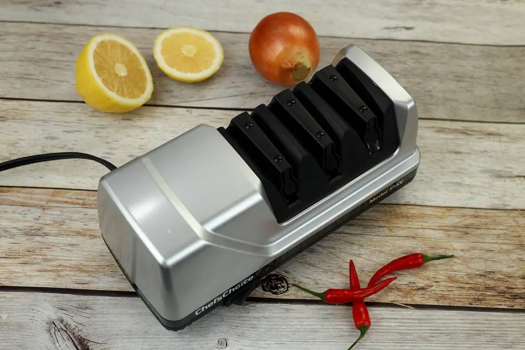 The Chef’s Choice Trizor XV electric knife sharpener on a wooden table surface with chili peppers, an onion, and two lemon slices for decoration.