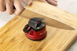 A hand holding a knife blade to sharpen on the Sunrise Pro manual sharpener, which stays on a cutting board.