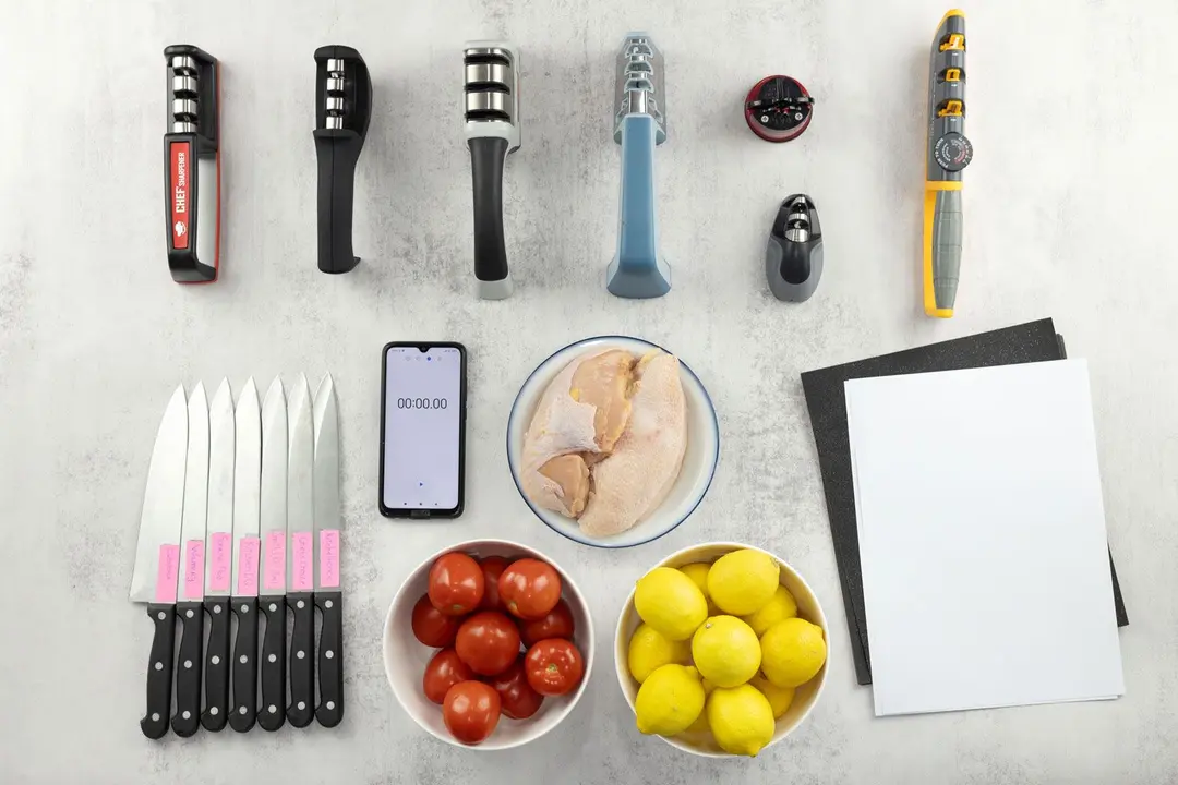 Pull-through sharpeners, including the Cubikook, Kitchellence, among others, plus testing equipment including test knives, one bowl of tomatoes, one bowl of lemons, one plate with raw chicken breasts, sandpaper, print paper, and a smartphone.