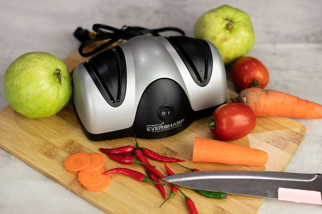 The Presto 08800 EverSharp electric knife sharpener on a cutting board with a knife and chilli peppers, guavas, carrots, tomatoes for decoration