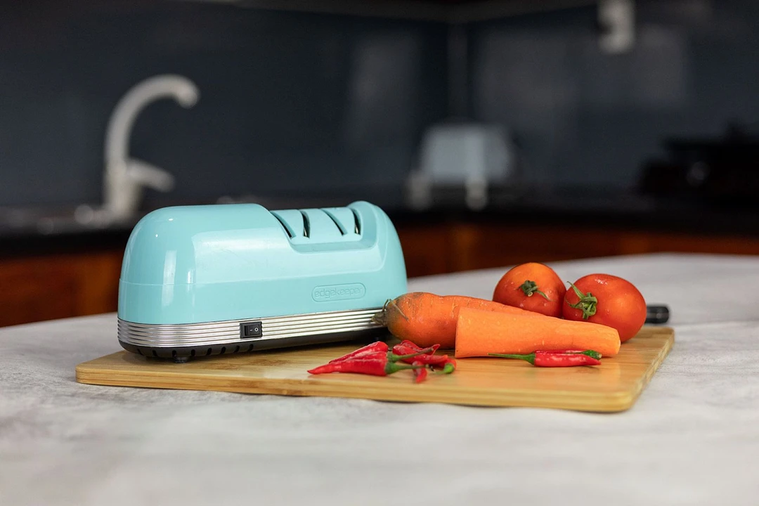 e EdgeKeeper electric knife sharpener on a cutting board with a chilli peppers, carrots, tomatoes for decoration and a faucet in the background