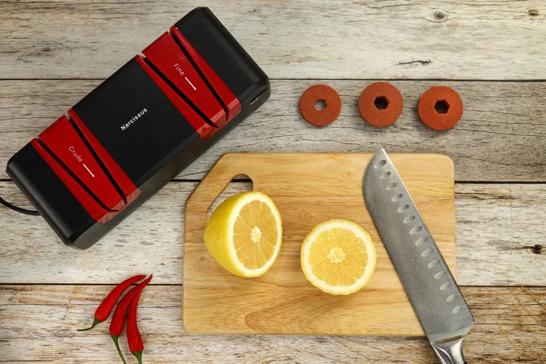 The Narcissus electric knife sharpener, three spare wheels, chilli peppers, a cutting board with a knife and slices of lemon.