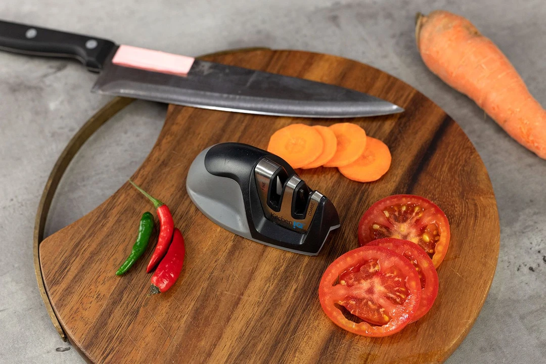 The KitcheniQ Edge-grip knife sharpener on a cutting board, kitchen knife, chilli peppers, carrot, tomato slices