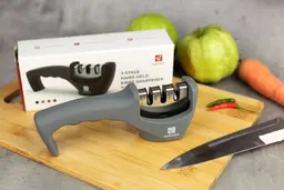 The Amesser 3-stage manual knife sharpener on a cutting board, its package box, knife, chilli peppers, carrot, guavas
