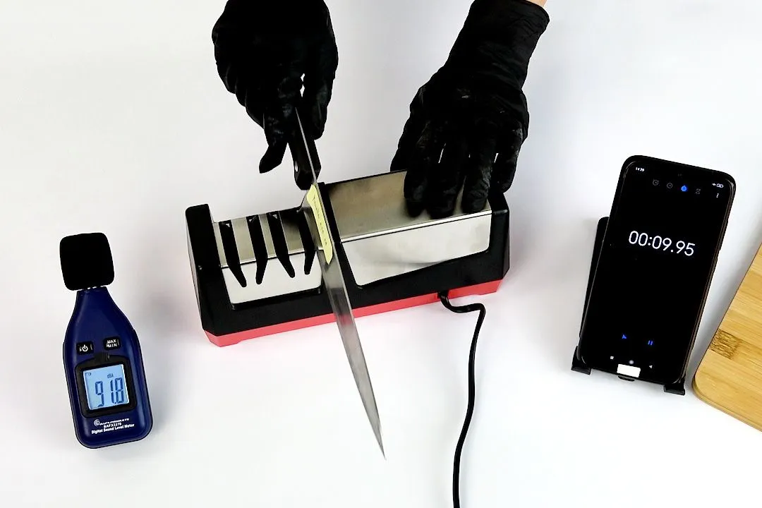 kitchen knife on Mueller electric sharpener, two hands in black gloves, smartphone timer, decibel meter, corner of cutting board