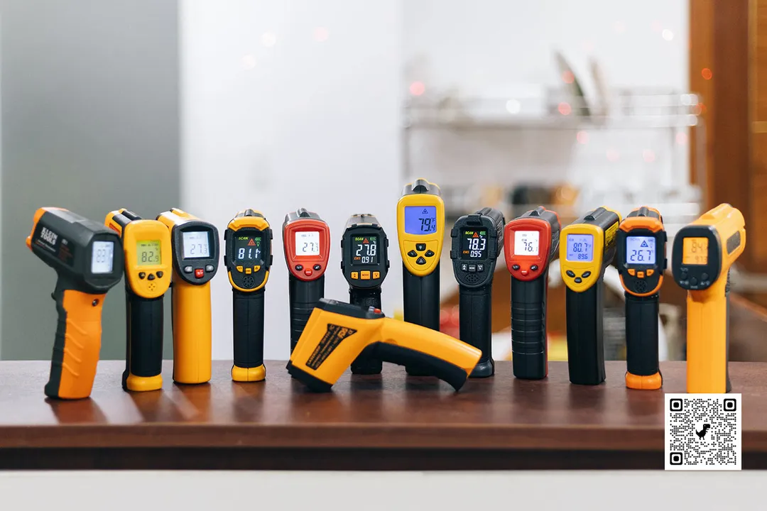 Thirteen best infrared thermometers in 2024 arranged in a line-up on a wooden countertop, against a blurry kitchen background.