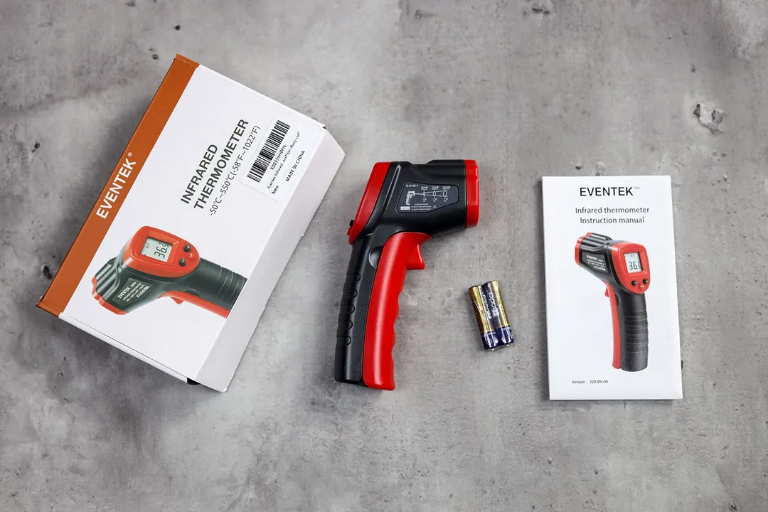 The Eventek IR thermometer in the center of the frame on its side. To the left is the box, and to the right is the battery and user manual.