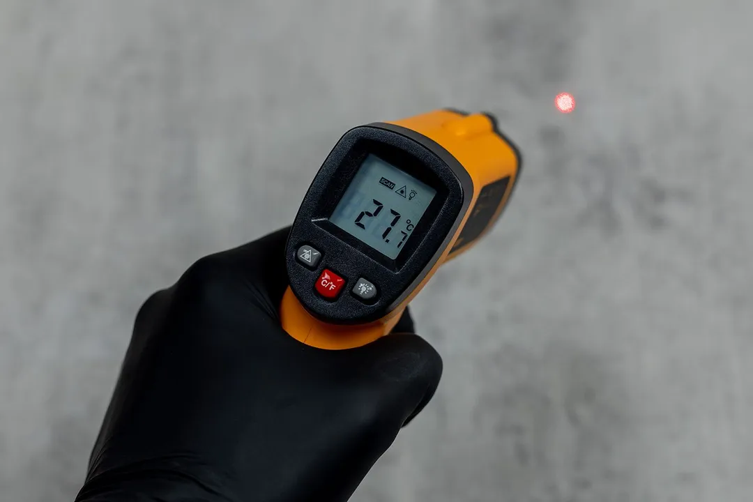 The Helect IR thermometer in the black-gloved hand of a reviewer. The thermometer is in scanning mode and there’s a laser dot in the background.