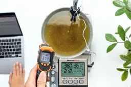 A reviewer is using the Kizen LaserPro LP300 at a distance of 16 inches to measure the surface temperature of a pan of hot oil. The screen reads 374°F.