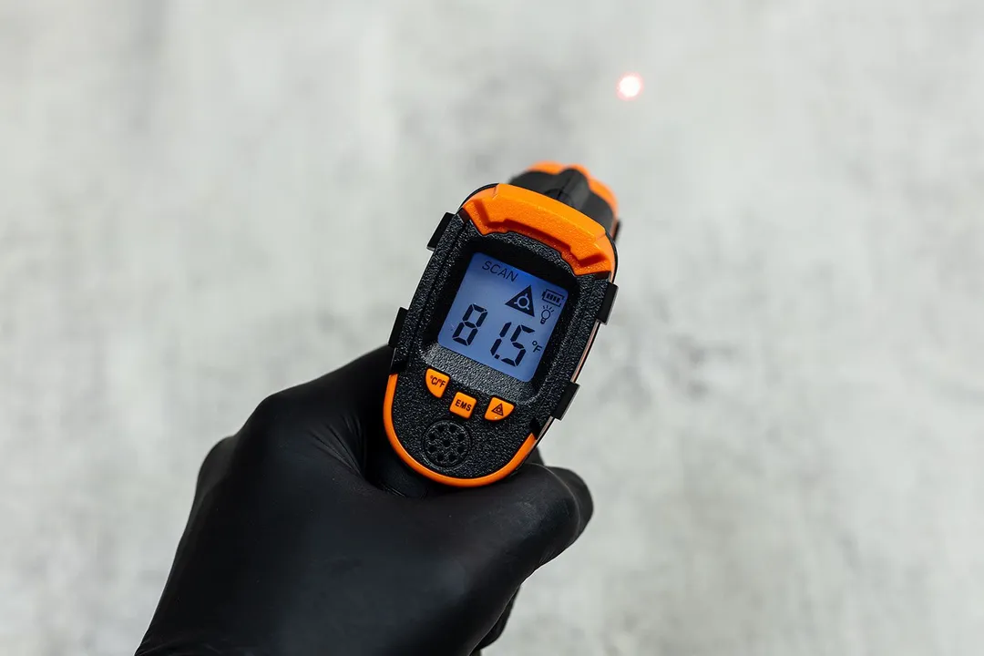 A black-gloved hand is holding the Kizen LaserPro LP300 IR thermometer and scanning the temperature of a white table top.