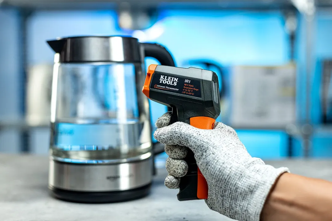 Klein Tools IR1 Infrared Thermometer in the gloved hand of a reviewer against a blurry blue backdrop with a kettle.