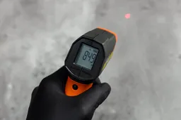 The black-gloved hand of a reviewer holding and using the Klein Tools IR1 IR thermometer in scan mode. A red laser dot can be seen in the background.