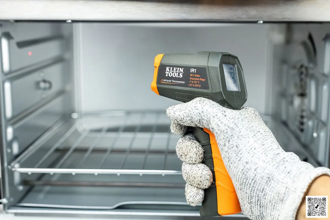A reviewer wearing white gloves is holding and using the black-and-orange Klein Tools IR1 IR thermometer to check the internal temperature of a toaster oven.